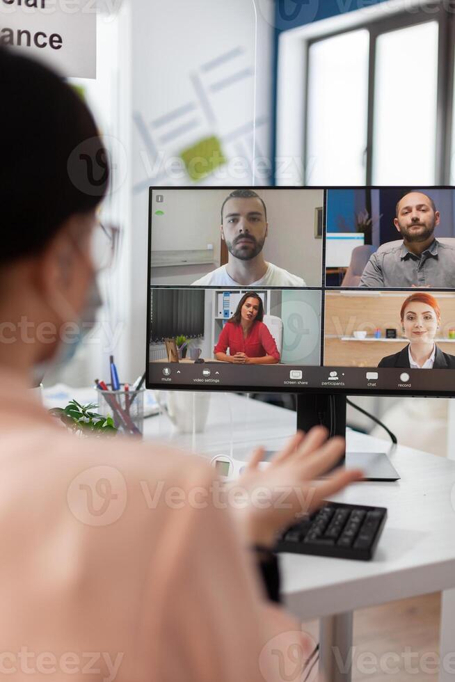 Executive manager with medical face mask discussing management statistics with remote team having online videocall meeting conference on laptop working in startup office. Teleconference on screen photo