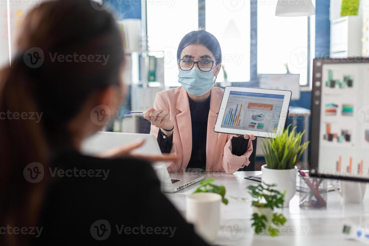 Businesswoman wearing medical face mask against covid19 showing financial graph to collegue using tablet computer working in startup office. Teamwork planning marketing strategy during global pandemic photo