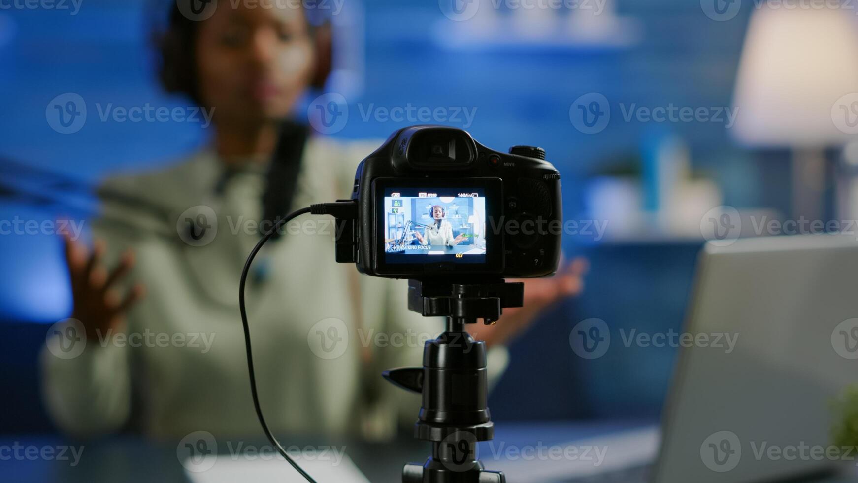 Close up of professional camera sitting in front of dslr recording online show. African woman vlogger speaking at microphone during livestreaming, blogger discussing in podcast wearing headphones photo