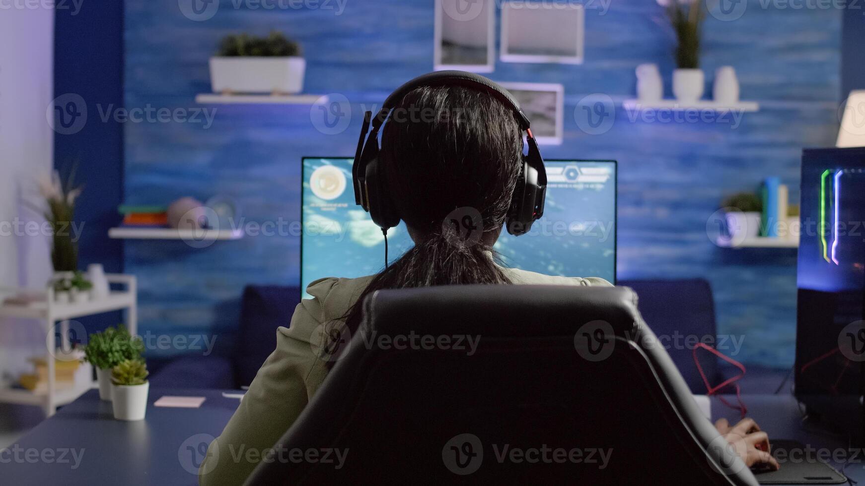 Back shot of streamer woman playing on RGB powerful computer shooter game for tournament, talking with multiple players into headphones. Pro gamer using professional streaming equipment photo