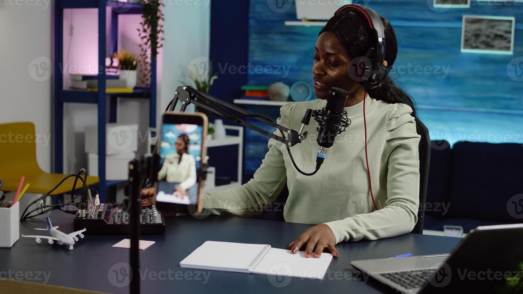 Smiling influencer african woman sitting in front of phone camera recording for blog. Digital blogger vlogger streaming talk show in studio using headphones, professional podcast microphone photo