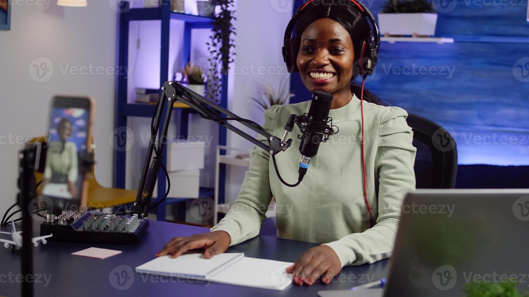 African creator content waving at camera smiling and talking with audience during podcast using smartphone. Influencer looking at phone while speaking at podcast microphone sitting in home studio photo
