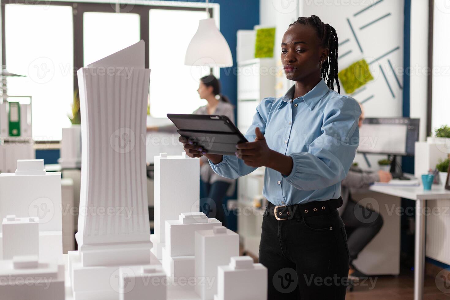 Architect woman of african american ethnicity working on tablet looking at professional maquette building model. Architectural worker viewing design for modern project in development photo