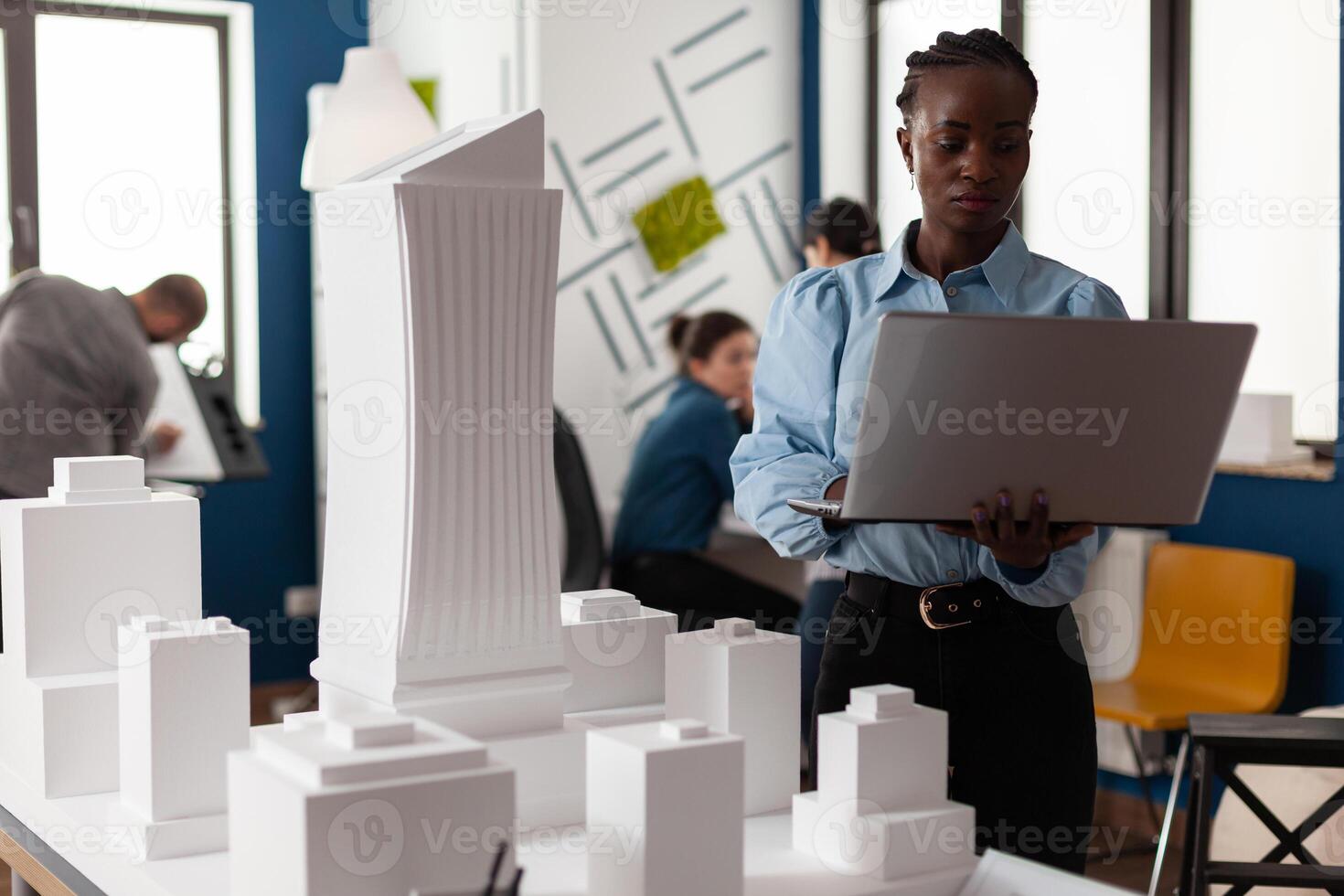African american architect at business workplace looking at laptop and blueprint plan for building model maquette. Engineer constructor working on modern layout design for project photo