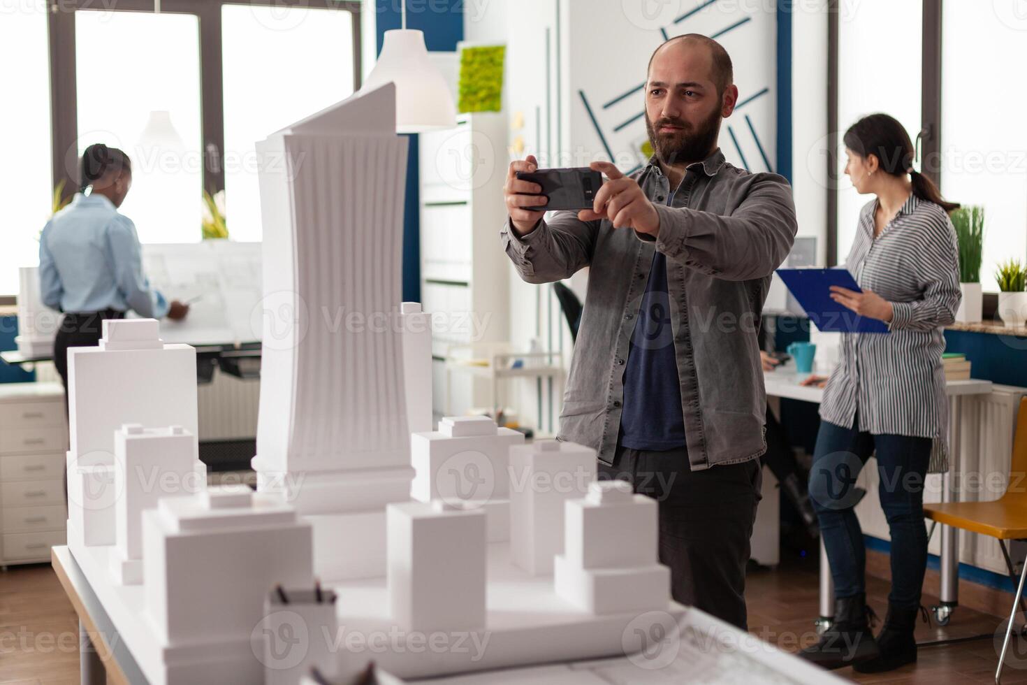 Design constructor looking at smartphone for project of architectural structure. Engineer standing at desk inspecting building model maquette urban plan layout for modern development photo