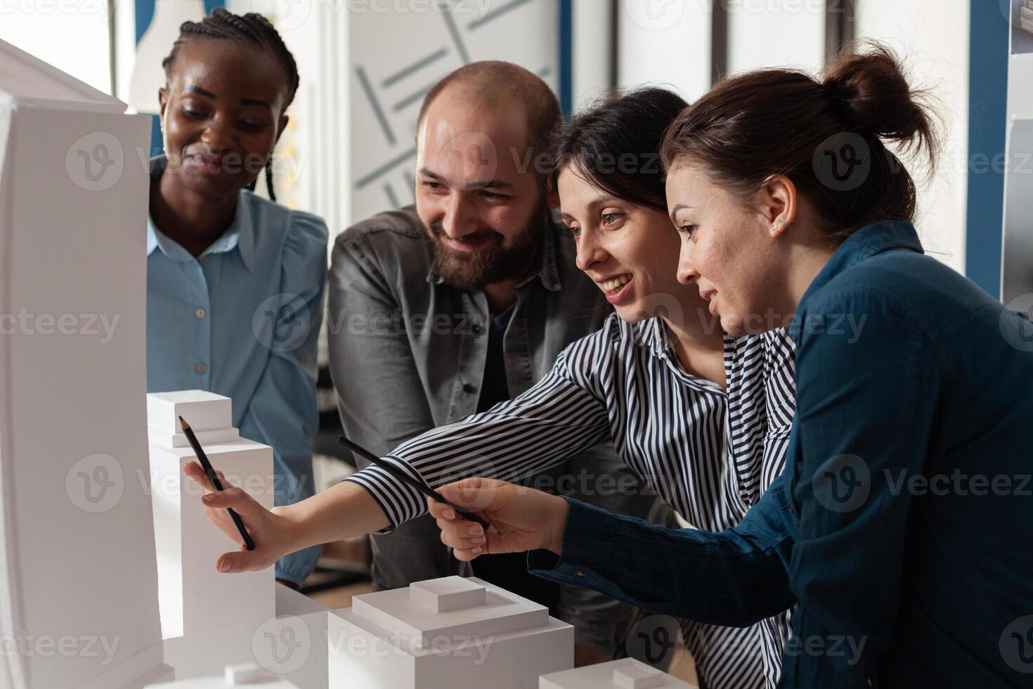 Multi ethnic architectural teamwork analyzing maquette building model construction layout. Diverse professional group of colleagues pointing at project design blueprint plan photo