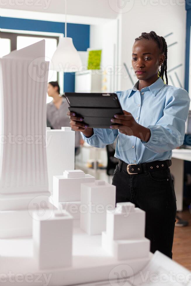Portrait of architect working on tablet standing next to architectural maquette of office buildings construction complex. Engineer comparing blueprints on digital device with scale foam model. photo