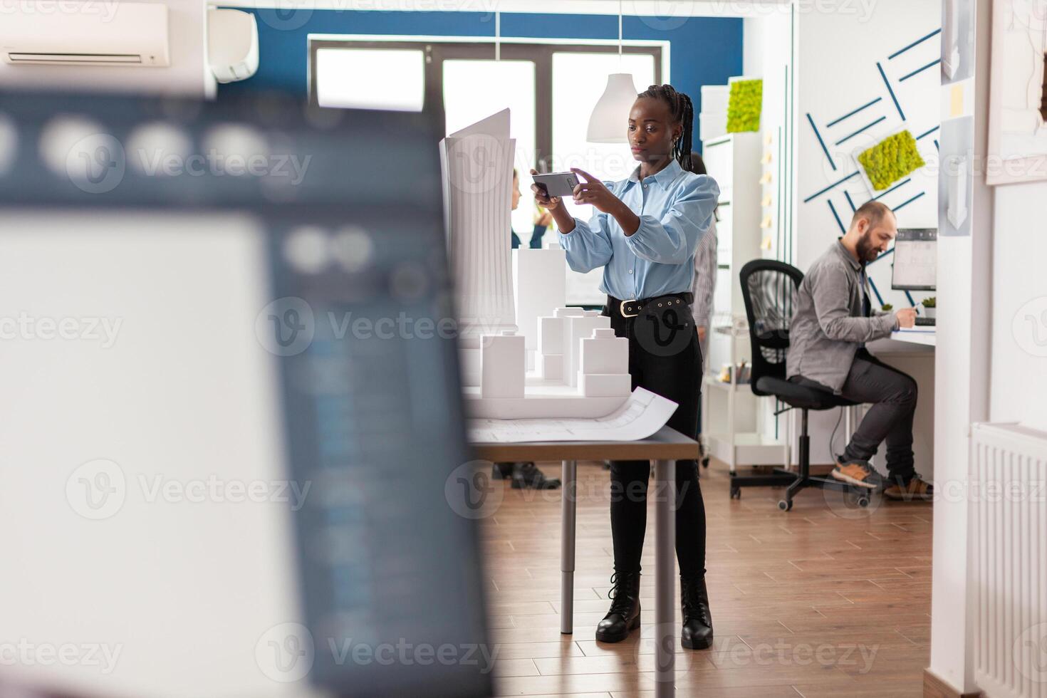 Architect working with smartphone standing next to table with white foam scale model in architectural modern office. Engineer with mobile device photographing maquette of real estate project. photo