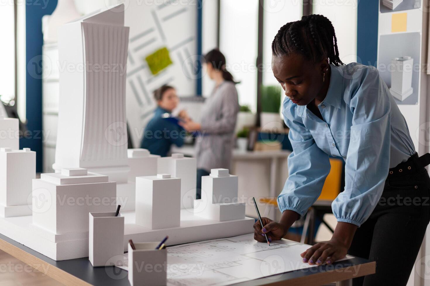 Residential architect drawing blueprints at desk in architectural office next to white foam scale model of skyscraper. Development engineer making notes on construction plan. photo