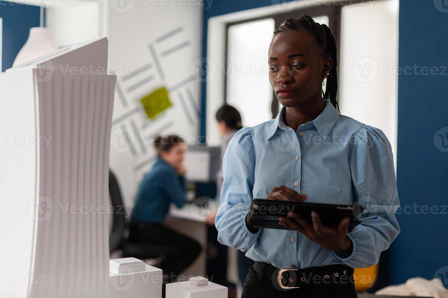 African america architect looking at scale foam model of modern building while holding tablet with blueprints. Architectural engineer focused on the design of 3d skyscraper maquette. photo