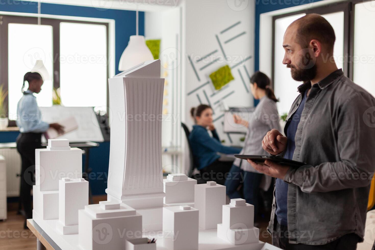 Architect holding tablet with blueprints inspecting design of white foam building model in architectural office. Engineer comparing construction plans with skyscraper maquette. photo