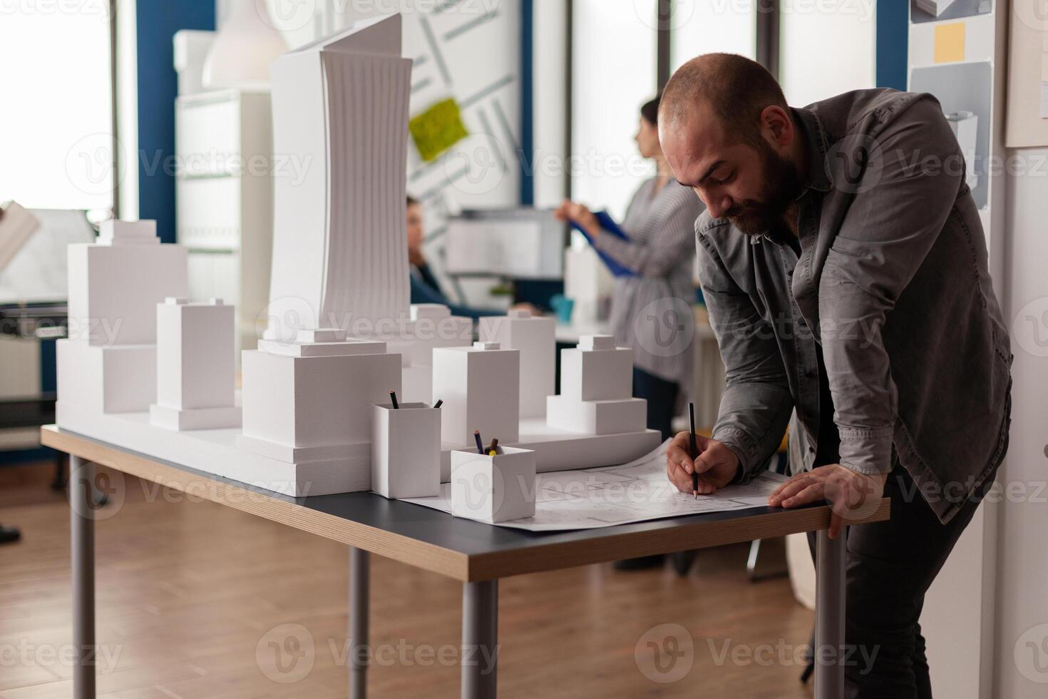 Development engineer making notes on construction plan of residential project leaning over desk with maquette. Architect drawing blueprints in architectural office next to white foam scale model. photo