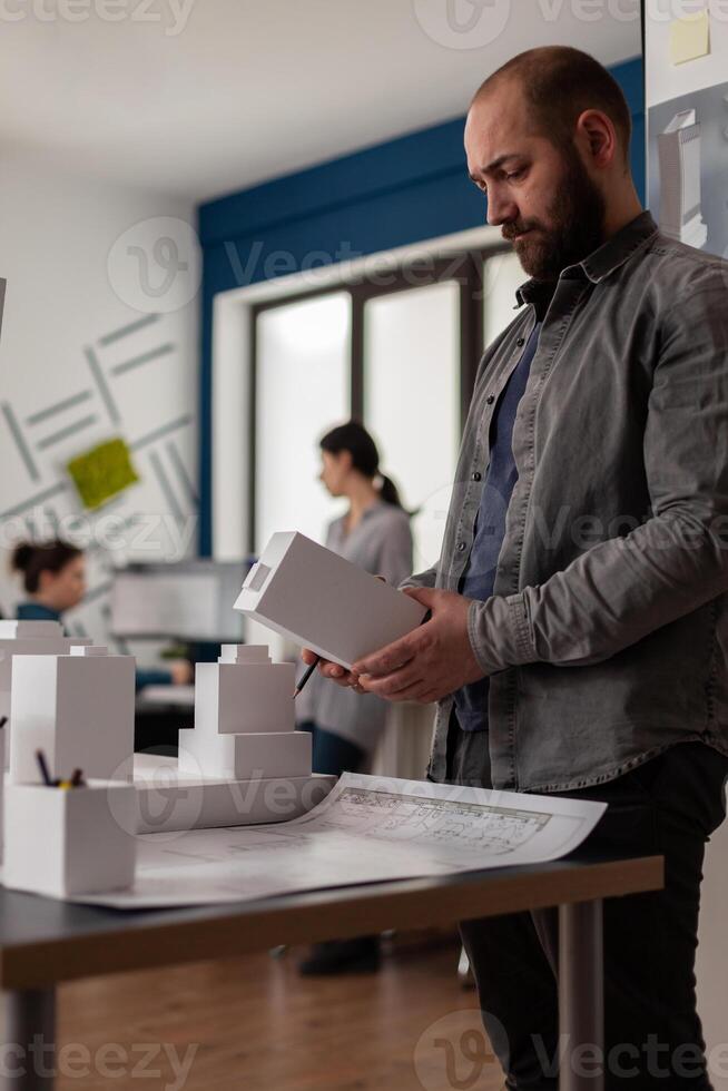 Building architect holding foam machette in hands looking for inspiration standing next to desk with blueprints. Architectural engineer in modern office comparing construction plans with 3d model. photo