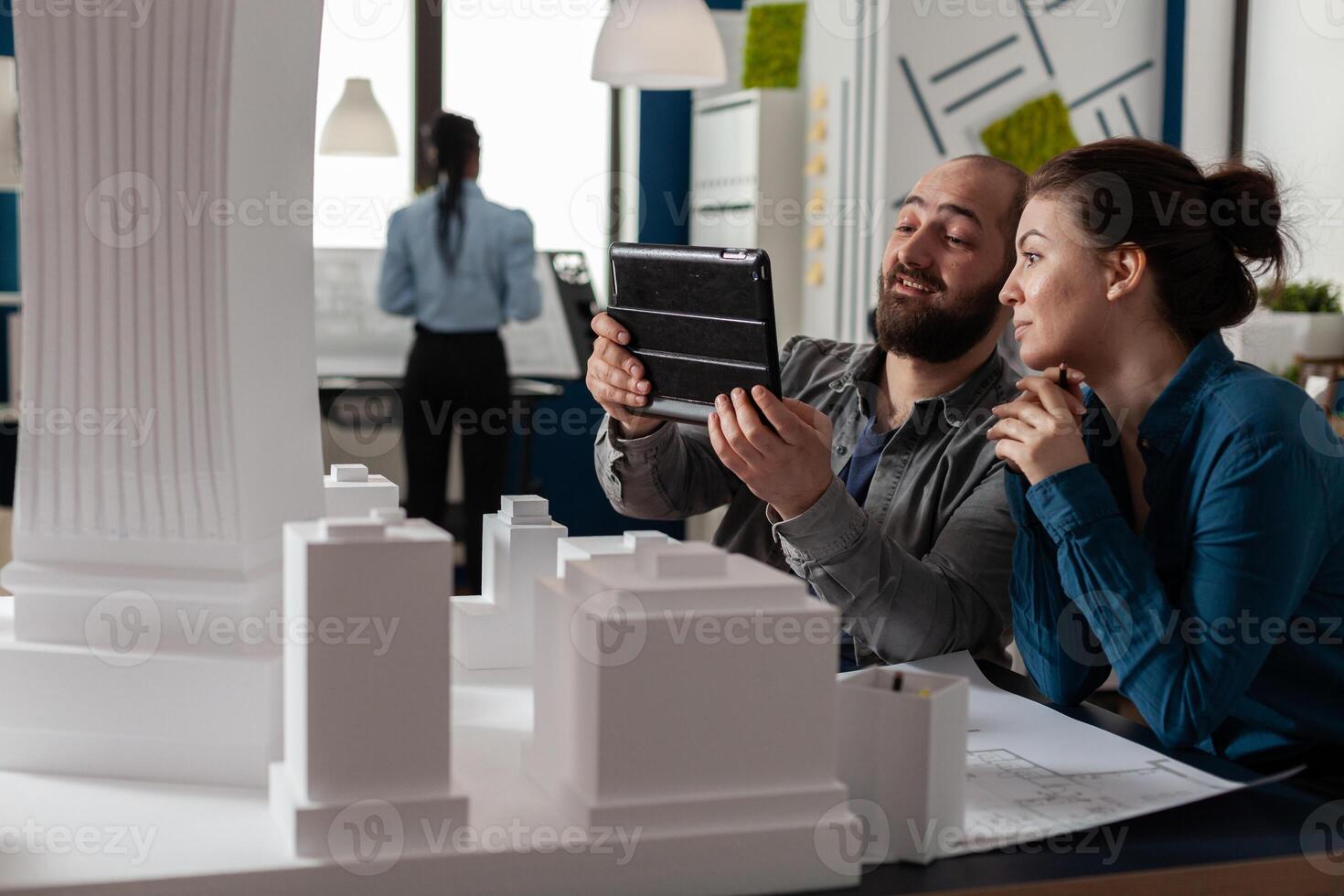 Two architects working together looking at tablet with blueprints sitting at desk in modern architectural office. Project engineers doing teamwork using digital device next to table with scale model. photo