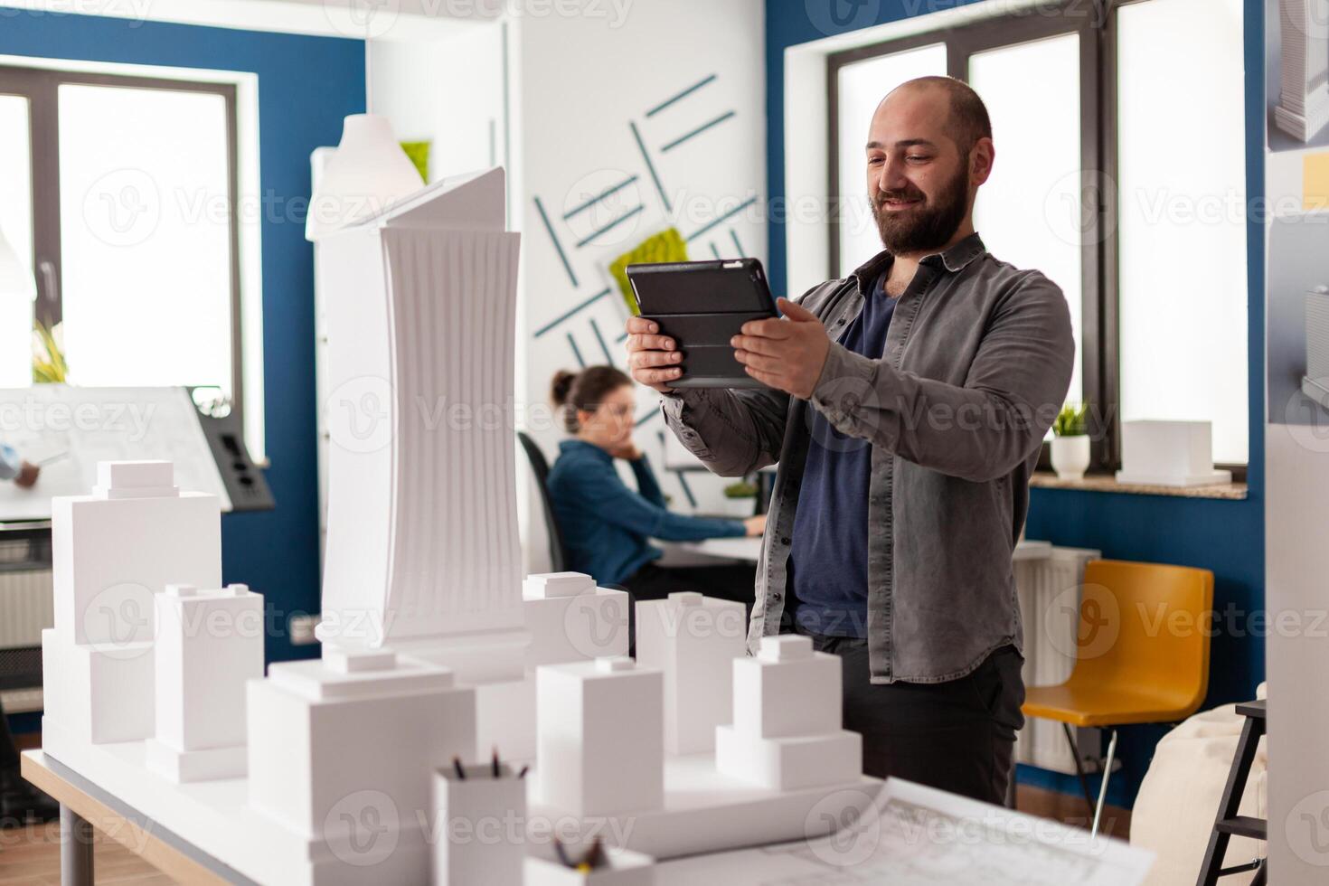 Bearded professional architect looking at tablet in front of white foam building model in architectural office. Engineer comparing blueprints on digital device with scale design of skyscraper. photo