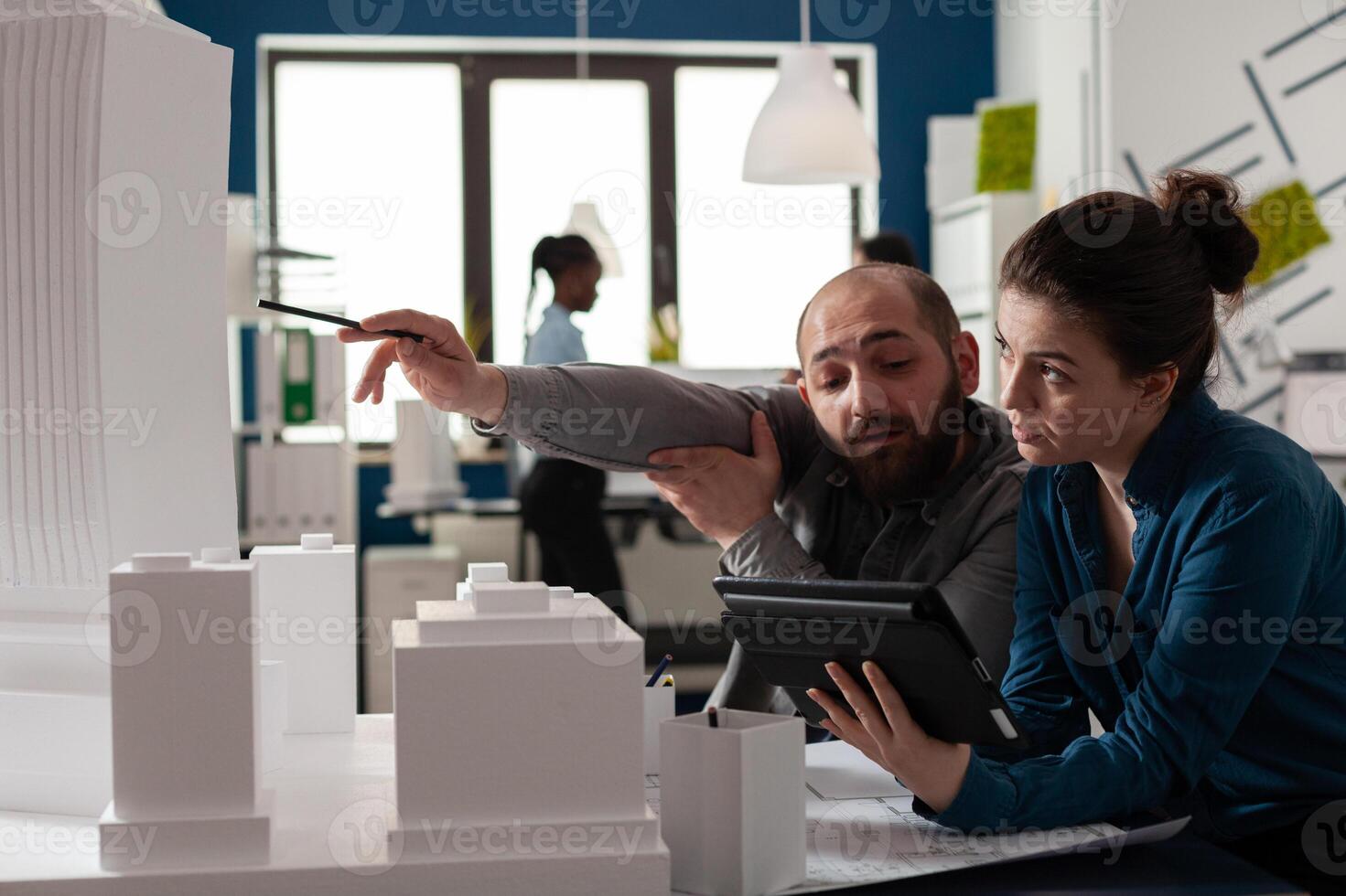 Architectural engineer pointing pen at skyscraper foam model collaborating with colleague engineer holding tablet with blueprints. Architects working together discussing building design. photo