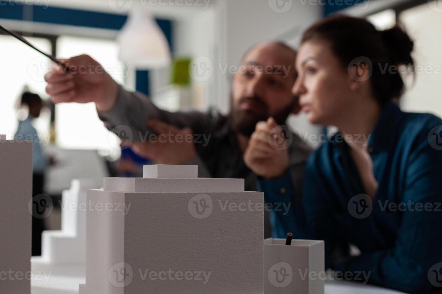 Closeup of white foam architectural scale model of buildings in front of architects team pointing at residential project. Detail of urban development maquette on design table. photo