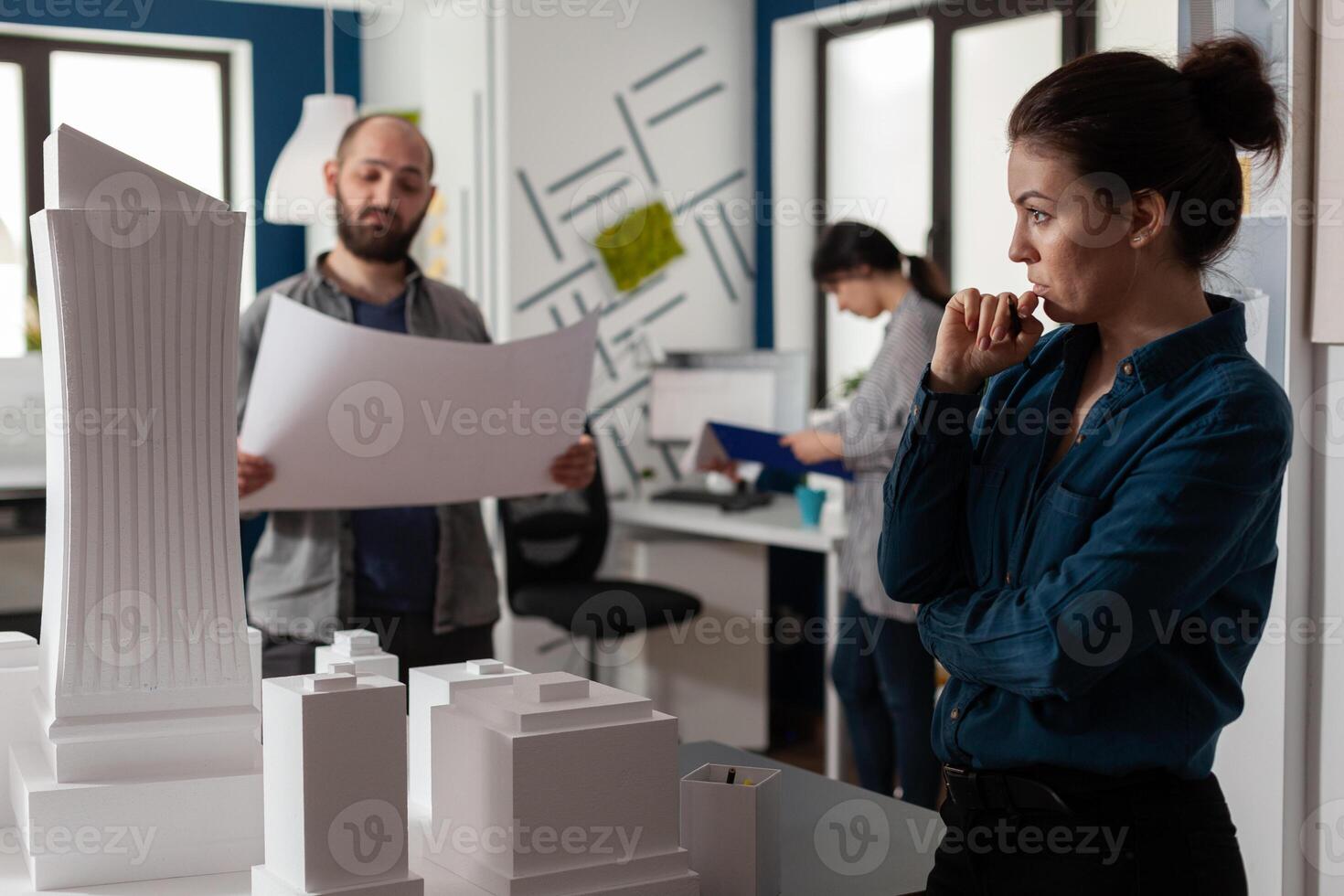 Architect looking into the distance gazing at skyscraper scale model with team colleague engineer holding construction plans. Team of architects brainstorming architectural ideeas. photo