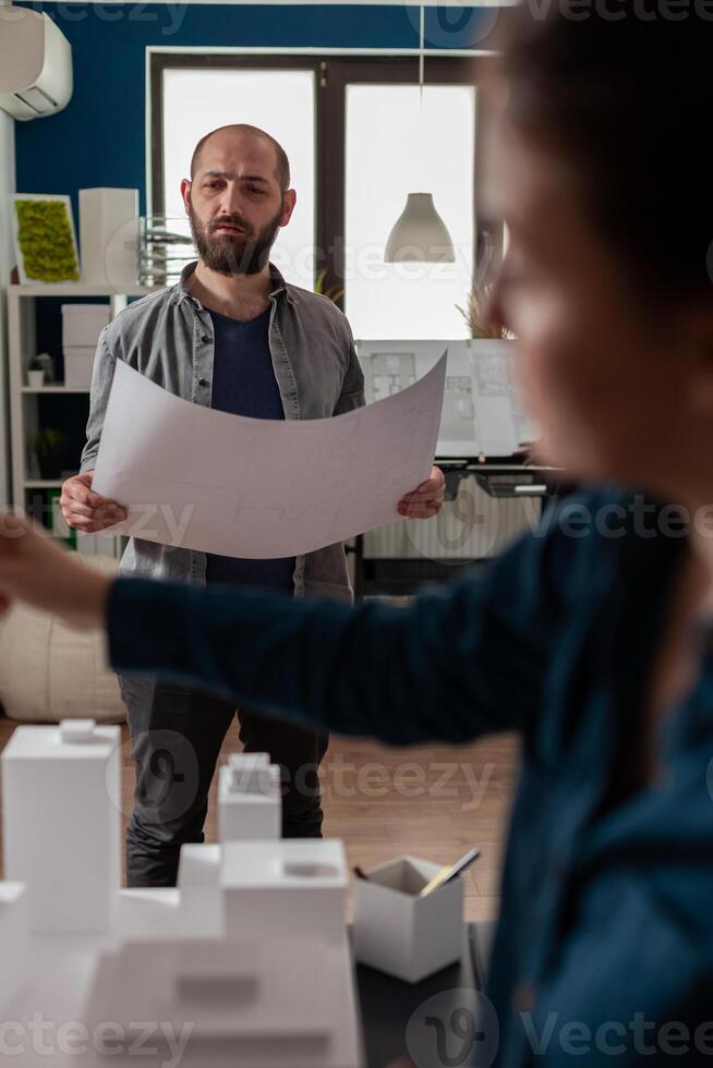Standing architect holding blueprints thinking about architectural design working together with colleague engineer in modern office. Architects doing creative teamwork on 3d buildings maquette. photo