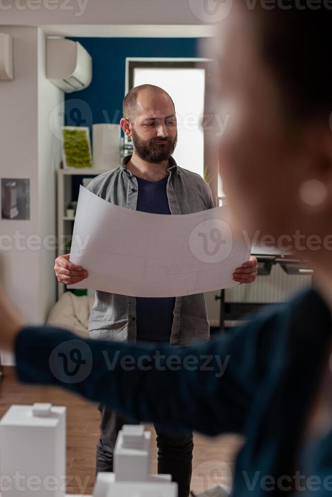 Standing bearded architect looking focused at construction plans collaborating with colleague engineer in modern office. Architects doing architectural design creative teamwork on foam scale model. photo