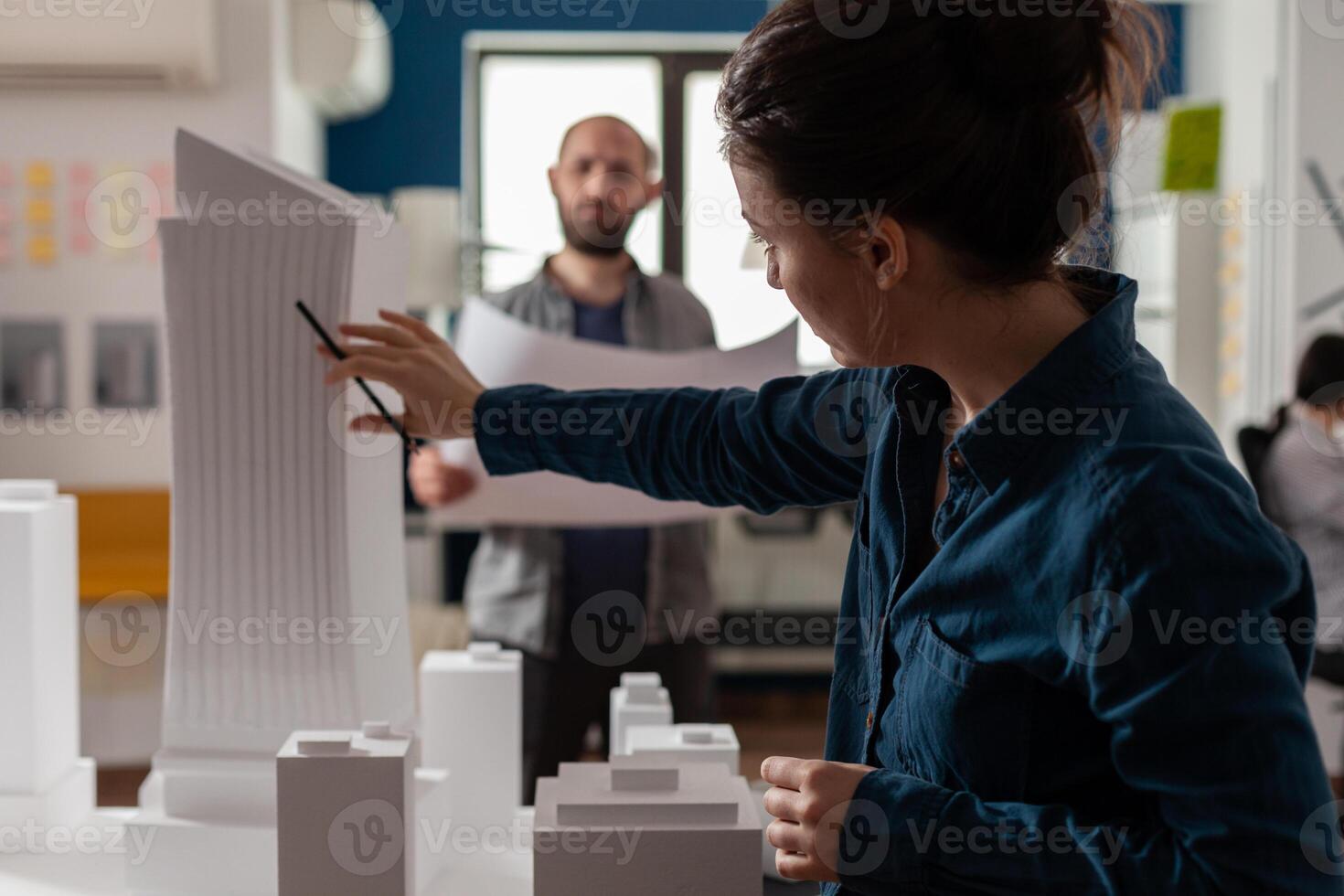 Architect inspecting futuristic building design of skyscraper with colleague engineer holding blueprints. Architects doing teamwork at desk with white foam maquette of construction project. photo
