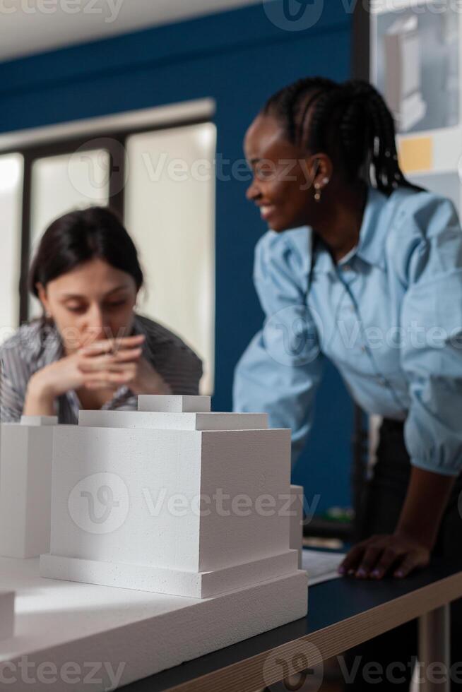 Closeup of white foam architectural scale model of buildings in front of architects team talking over blueprints. Detail of urban development maquette on design table. photo
