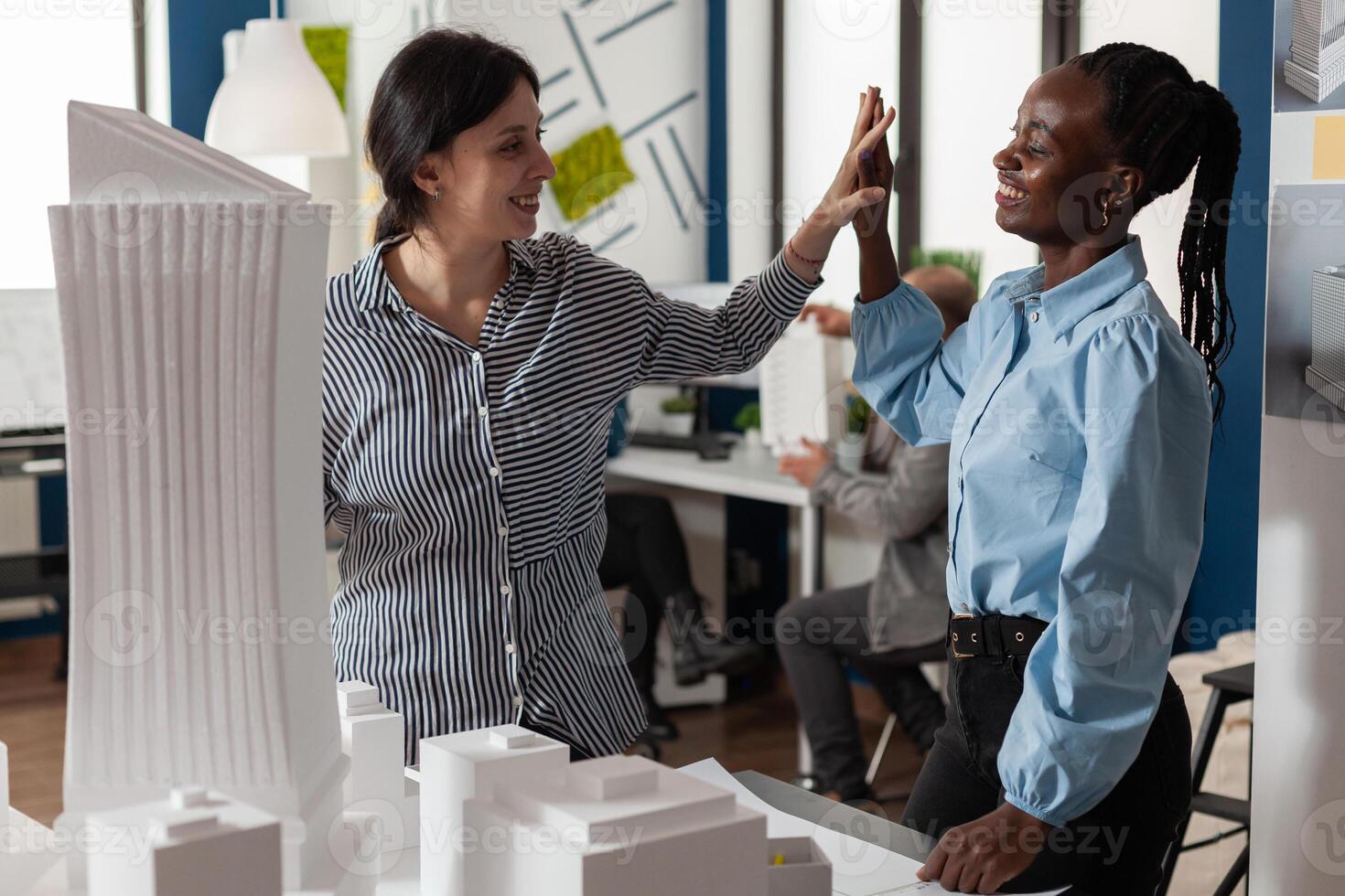 Team of two women architects high fiving standing in front of table with foam scale model of residential buildings in architectural modern office. Successful happy engineers celebrating team project. photo