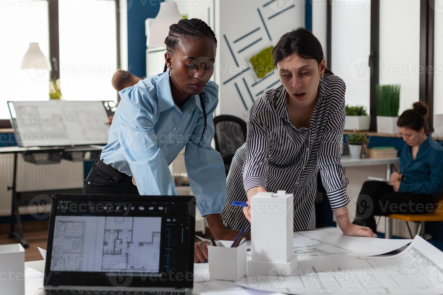 Team of two professional architects working on blueprints on desk in architectural office in front of laptop with construction plans. Colleagues discussing design of white foam building model. photo