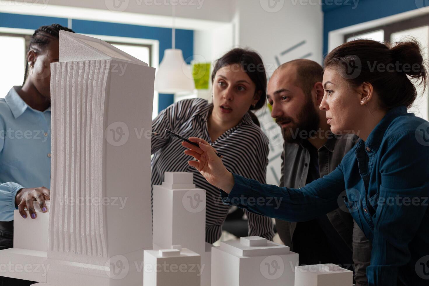 Professional architect with working partners pointing at skyscraper foam scale model in architectural office. Team of engineers collaborating on group project at desk with buildings maquette. photo