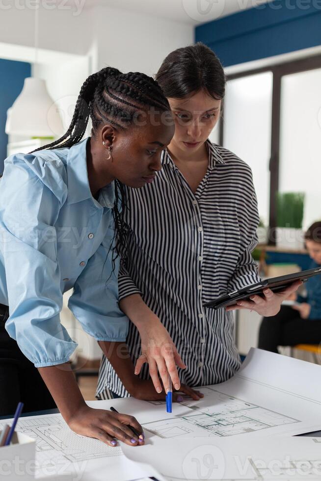 Architect holding digital tablet computer with construction plans explaining blueprints to african american engineer colleague. Professional architects in architectural office designing urban development project. photo