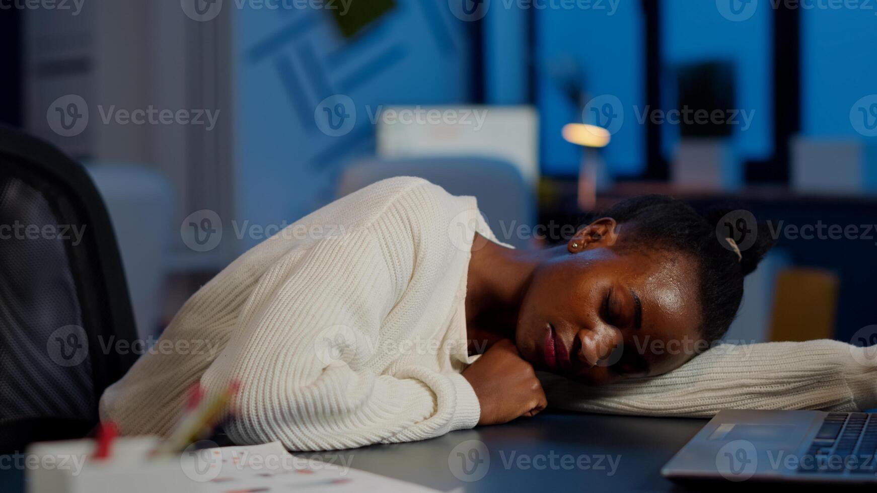 Dark skin freelancer doing overtime falling asleep with hand on desk trying to respect deadline for project. African employee working late at financial project sleeping on table in strat up office photo