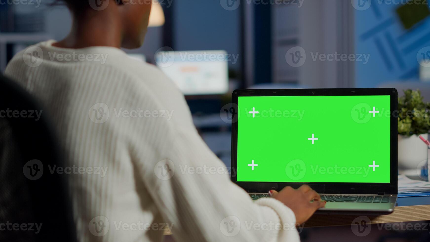 African woman manager looking at mock up monitor of laptop, green screen mockup, chroma key desktop sitting at desk in business office late at night. Freelancer watching at isolated desktop overwork photo