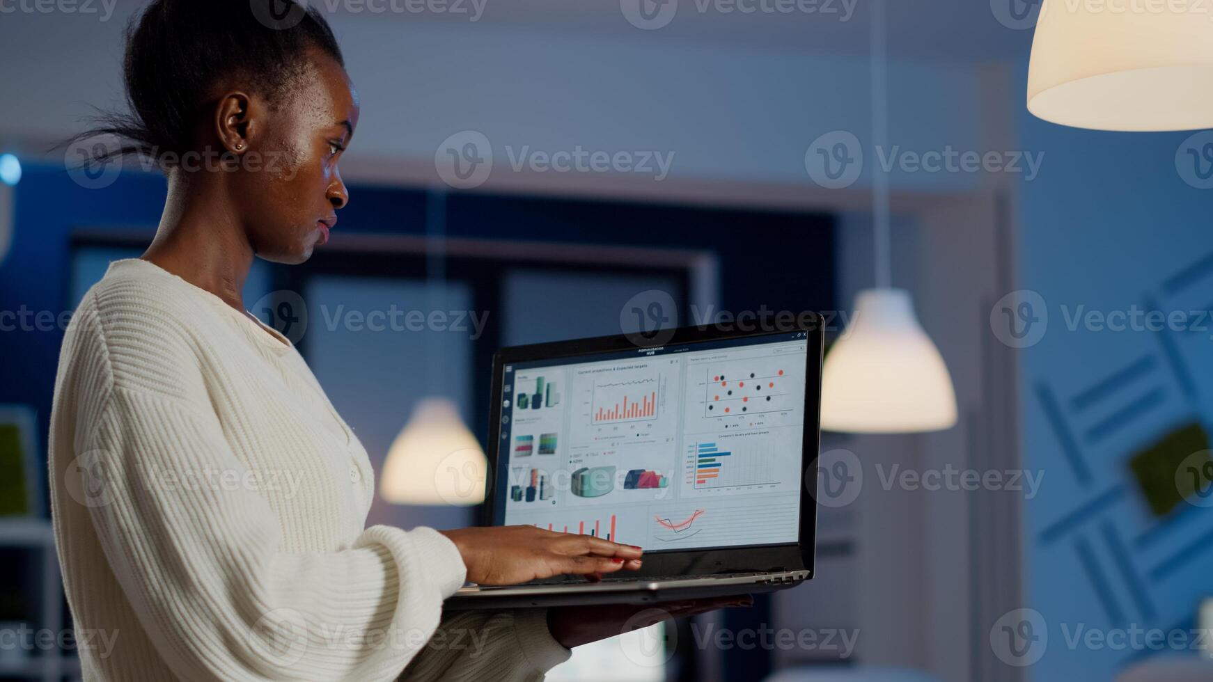 African freelancer typing on laptop late at night standing in start up business office late at night working. Busy employee using modern technology network wireless doing overtime for job searching photo