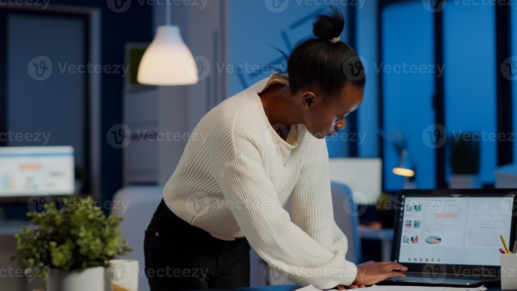 Stressed african manager woman working with financial documents checking graphs, holding papers, reading raports, standing at desk late at night in start-up office doing overtime to respect deadline. photo
