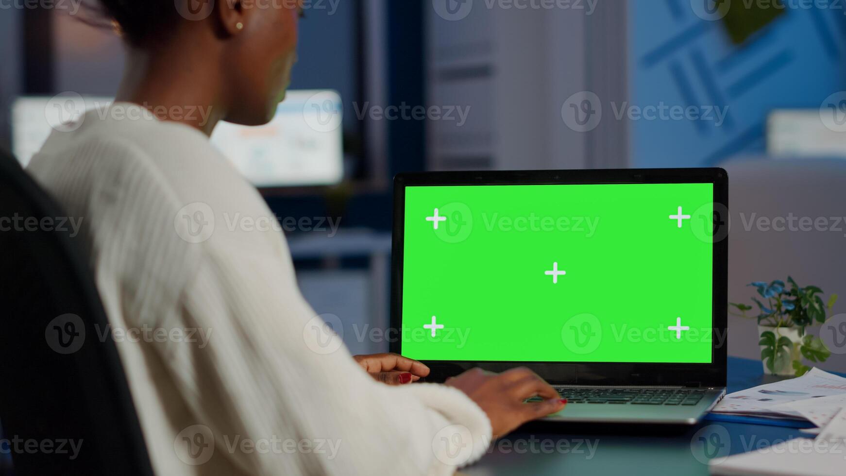 Black freelancer searching using laptop with greenscreen display, green mockup, chroma key desktop sitting at desk in business office working at night. Freelancer watching at isolated desktop overwork photo