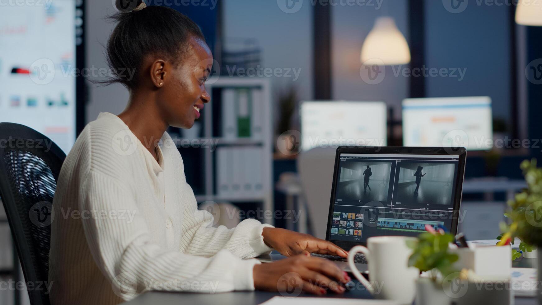 Black delighted videographer editing movie on professional laptop sitting at desk in business office at midnight. Creative editor working at night at new project processing audio film montage. photo