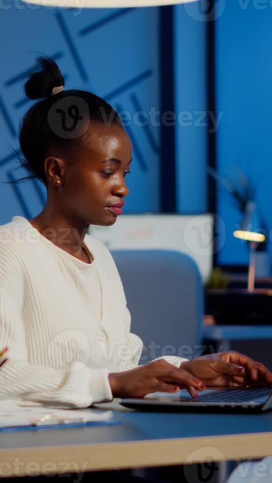 Busy african manager working on financial reports checking statistics graphs, typing on laptop sitting at desk late at night in start-up office doing overtime to respect deadline of financial project photo