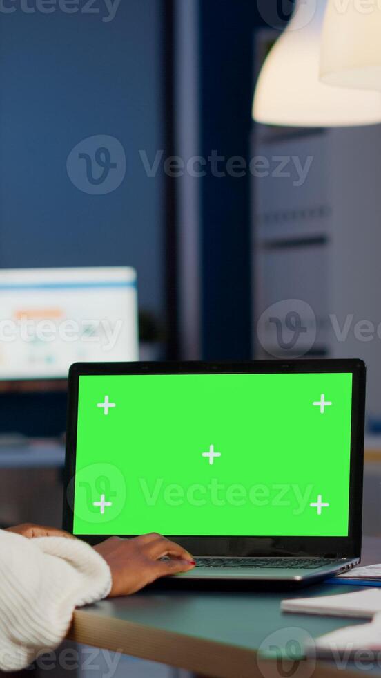 African manager typing at laptop with chroma key display during night time in start up business office working overtime. Freelancer watching mockup desktop monitor with green screen, writing datas photo