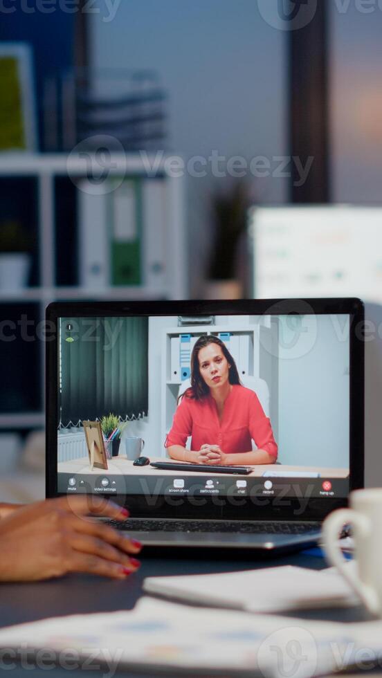 African businesswoman discussing with remotely woman partner online sitting in front of laptop working in start-up office talking on call during virtual meeting at midnight, using headphone photo