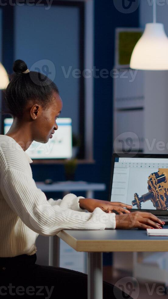 African engineer working late at night on a 3D model of industrial gears at laptop doing overtime in start up office. Freelancer studying prototype idea on pc showing cad software on device display photo