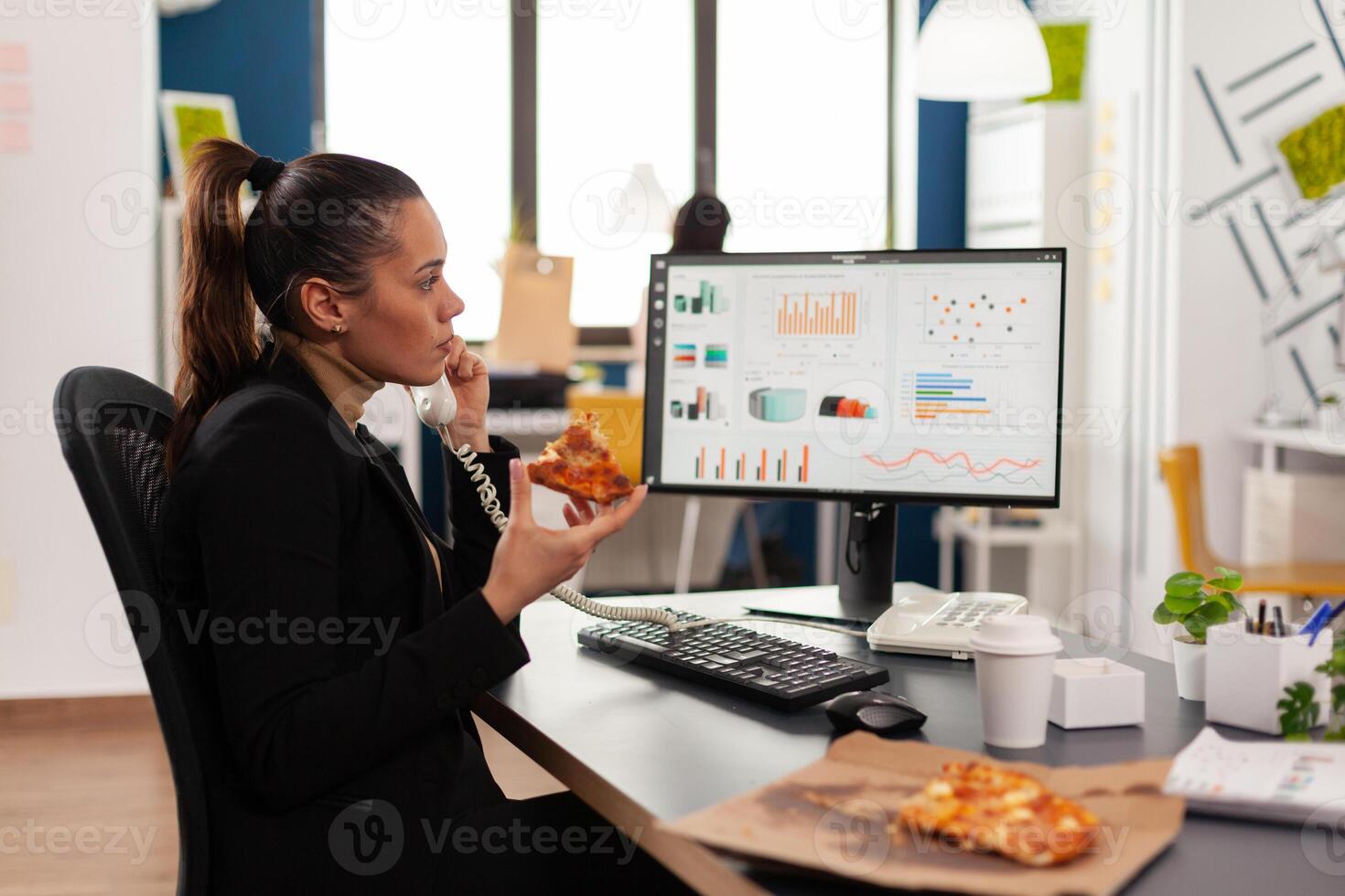 Closeup of businesswoman sitting at desk in front of computer eating pizza slice while talking at landline with remote company manager. Lunch meal break package delivered at startup office. photo