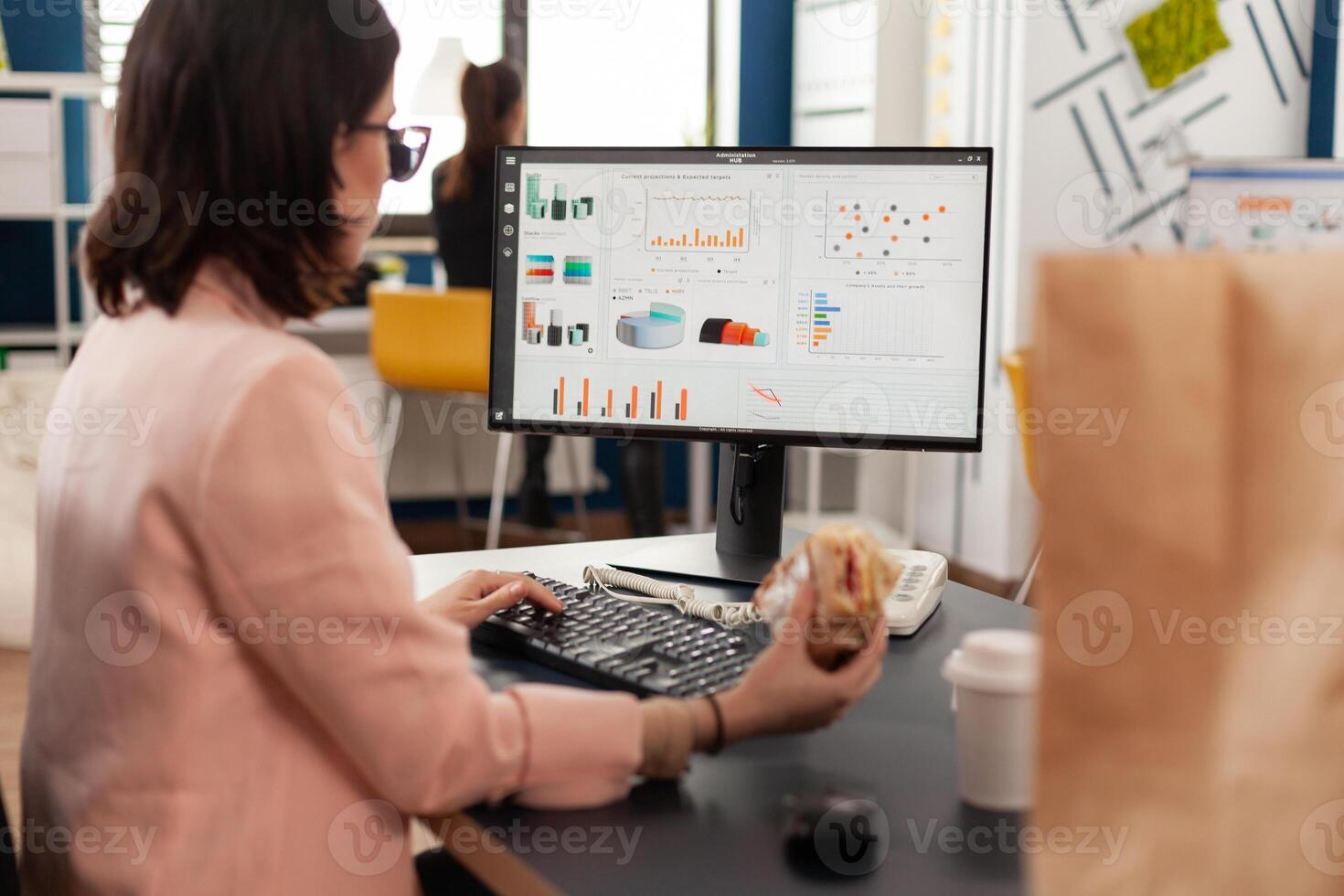 Businesswoman eating tasty sandwich having meal break working in business company office during takeout lunchbreak. Fast food order paper bag delivered at workplace. Woman typing strategy on computer. photo