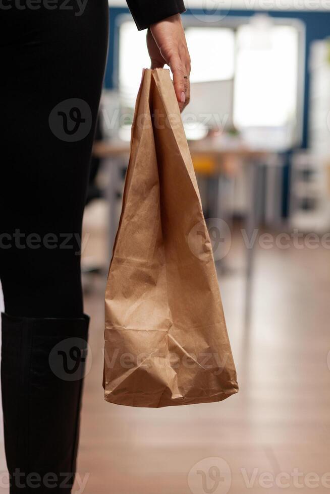 Businesswoman holding delivery takeaway food meal order paper bag during takeout lunchtime putting at desk in startup company office. Entrepreneur woman having lunch service while working at strategy photo