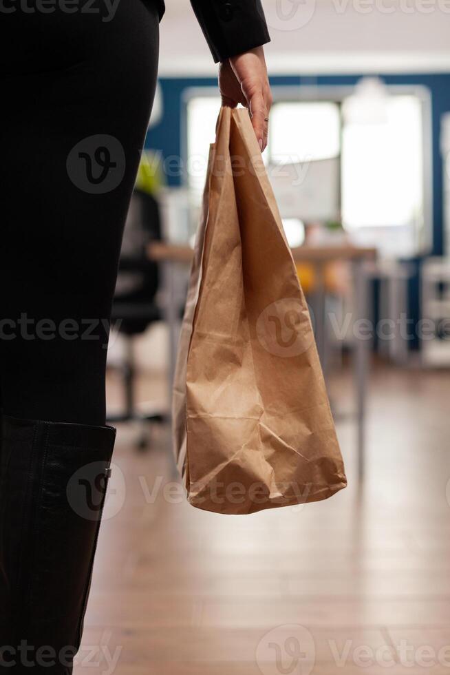 Businesswoman holding delivery takeaway food meal order paper bag during takeout lunchtime putting at desk in startup company office. Entrepreneur woman having lunch service while working at strategy photo
