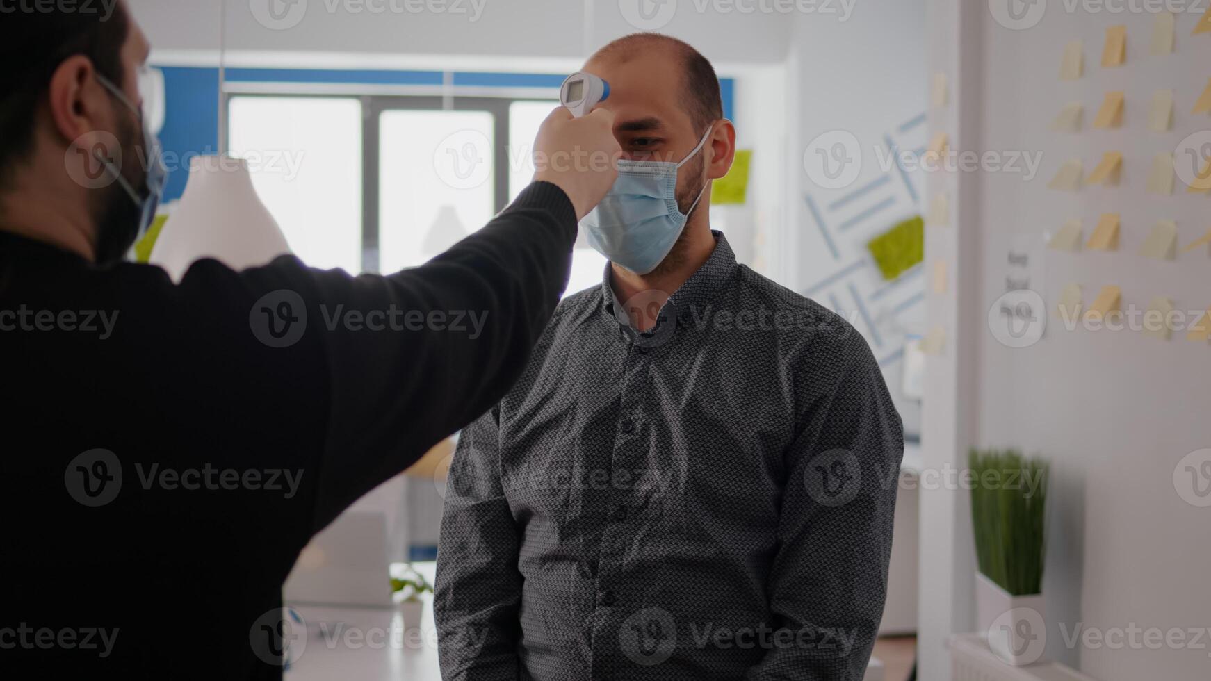 Freelancer man wearing protective mask against covid19 while checking temperature using medical thermometer to avoid infection with coronavirus. Company taking precautions during global pandemic photo