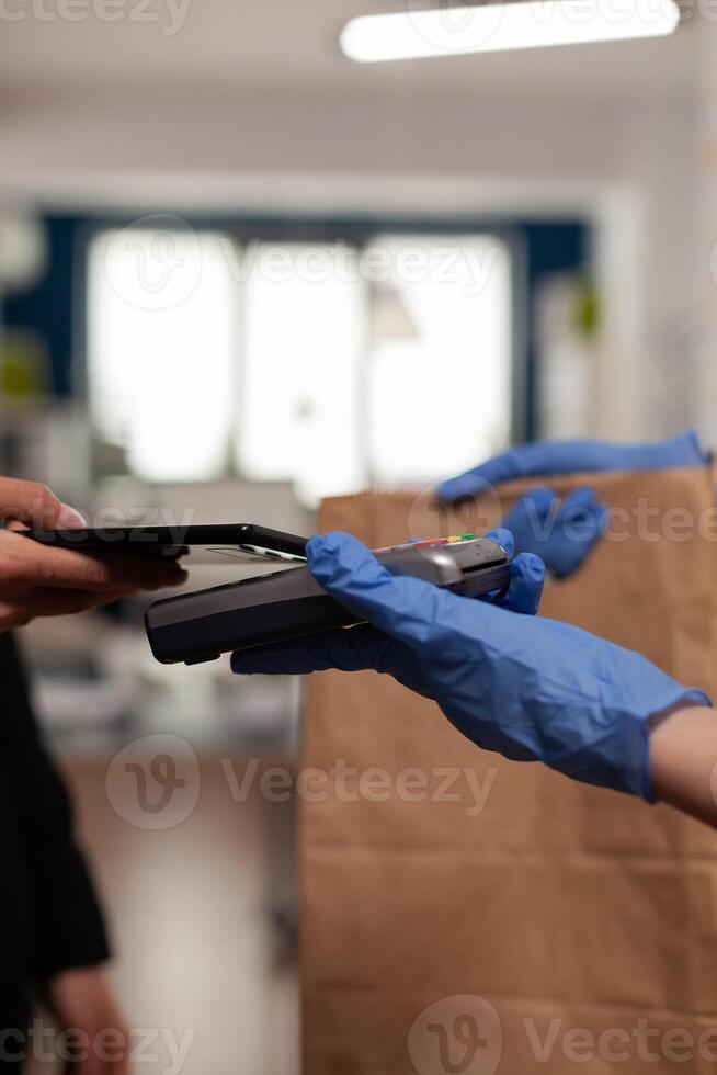 Delivery guy wearing protective gloves receiving paymant from businesswoman using smartphone nfc, technology over pos terminal service for delicious tasty meal lunch in paper bag. photo