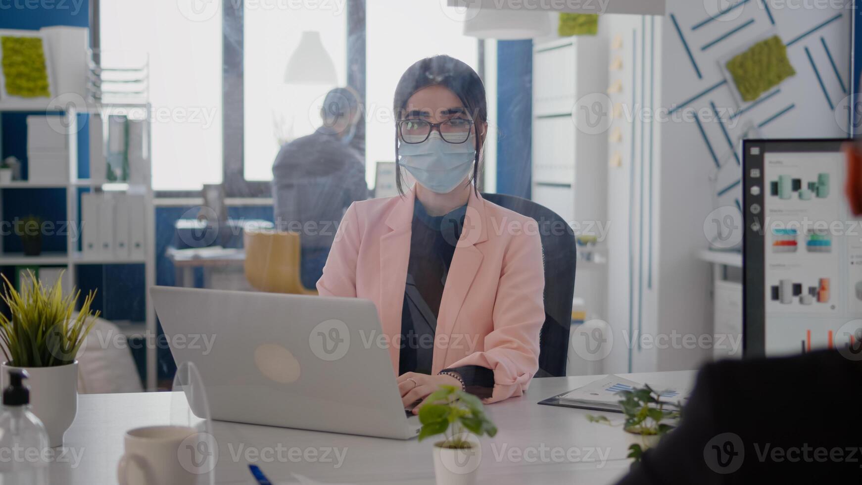 Portrait of business woman with protection face mask working on laptop computer in office during coronavirus pandemic. Coworkers keeping ing ing social distancing to prevent infection with covid19 virus photo
