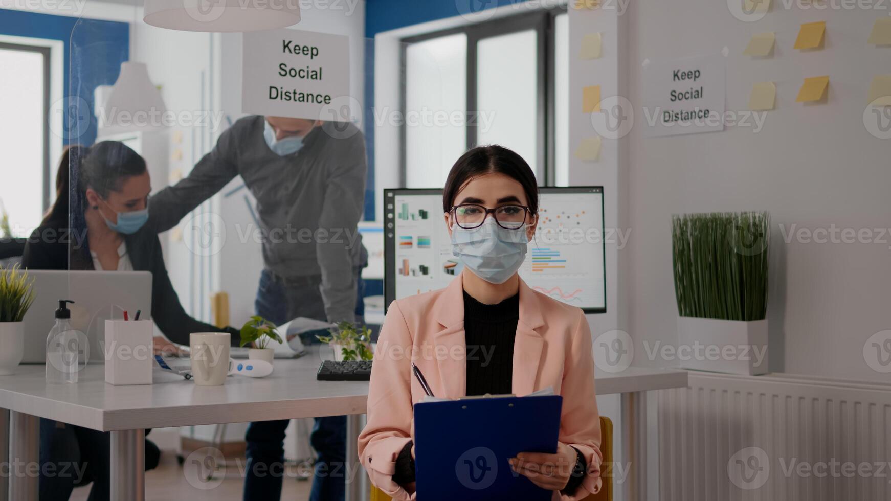 Entrepreneur sitting in new normal office looking into camera while writing lifestyle information during online videocall meeting. Businesswoman wearing face mask to avoid infection with covid19 photo