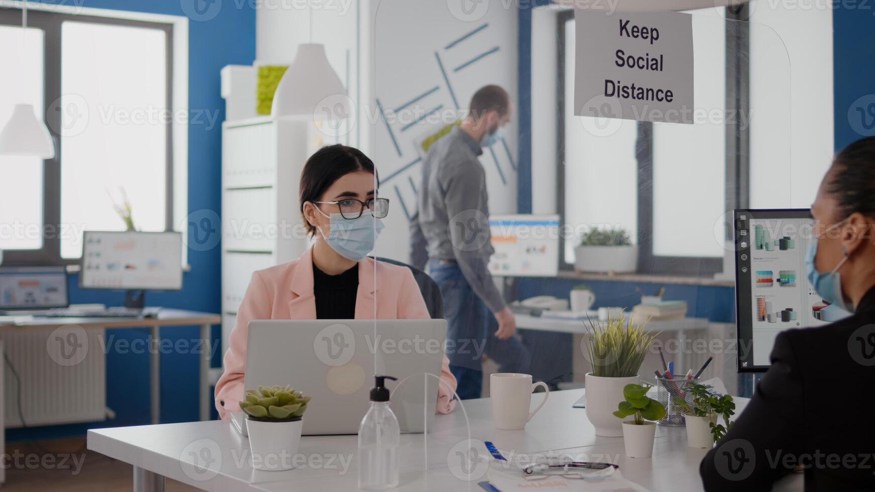 Business team with face masks planning marketing project while looking into monitor for statistics sitting in new normal office. Coworkers respecting social distancing during coronavirus global pandemic photo