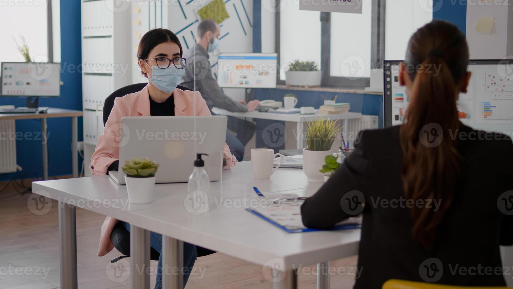 Freelancer talking with coworker about business startegy while sitting in new normal office wearing protective face mask to prevent infection with coronavirus. Team respecting social distancing photo